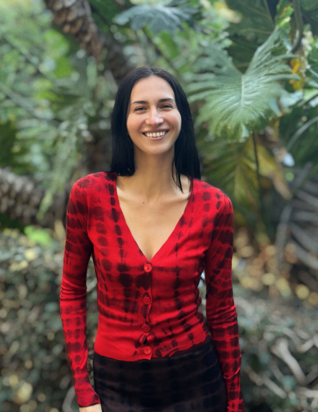 Women wearing a red cashmere tie dye cardigan in front of green foliage.